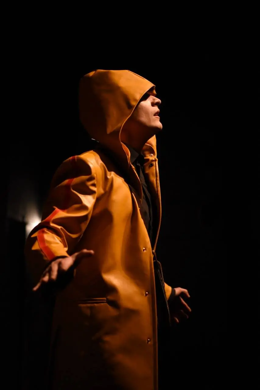 Une silhouette en manteau orange, de profil sur fond noir, éclairée latéralement — Antigone de Jean Anouilh, mise en scène d’Andréa Brusque.
