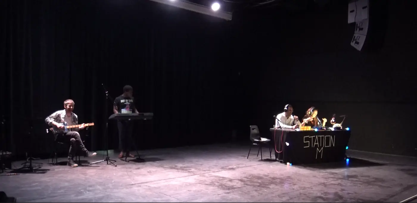 Sur le plateau, une table de mixage éclairée et des interprètes assis derrière un pupitre marqué « Station M ».